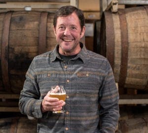 Man holding a beer standing in front of wooden barrels